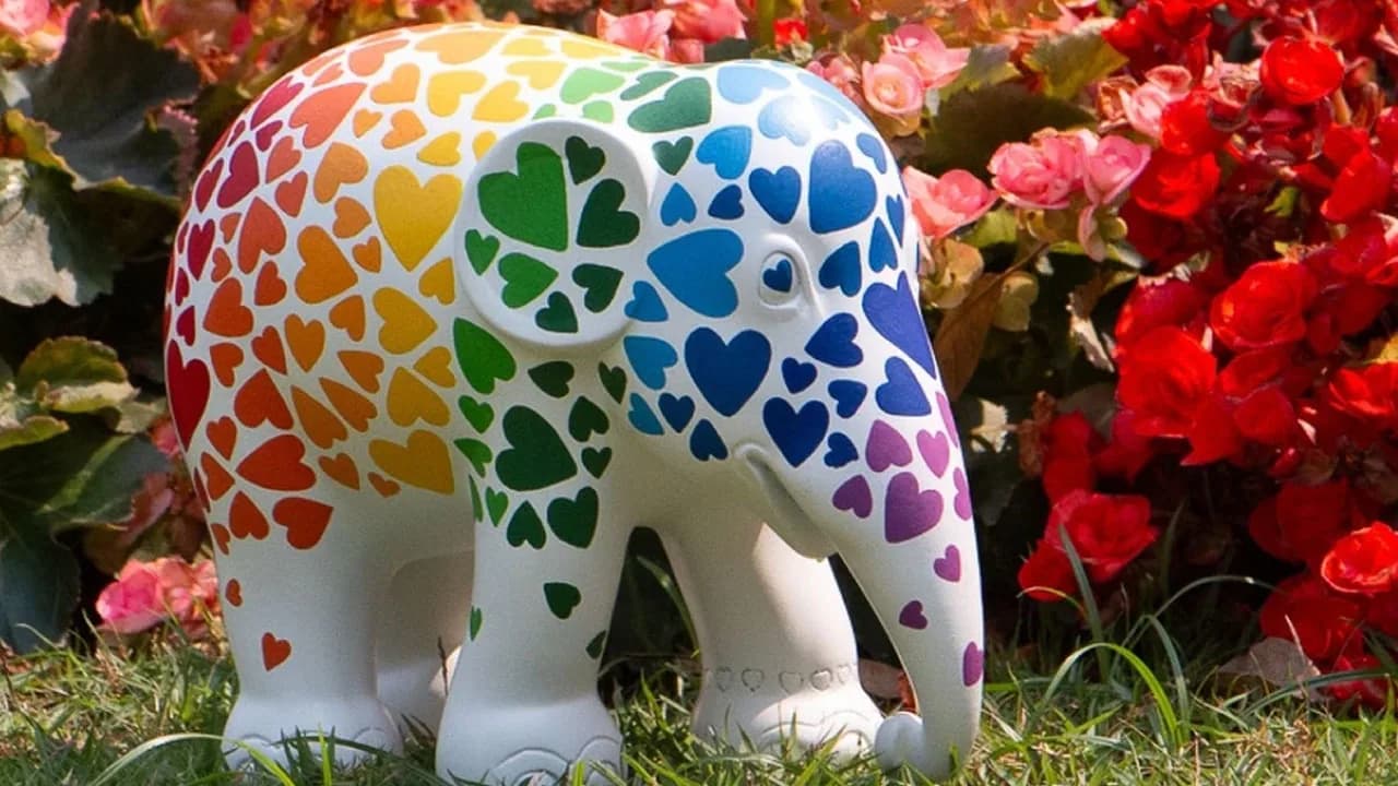 A white elephant sculpture painted with rainbow hearts, standing in red flowers, in Elephant Parade's launch image