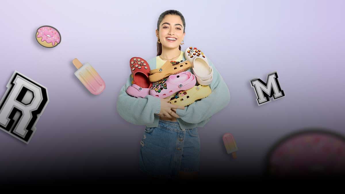 Rashmika Mandanna holding Crocs clogs and Jibbitz charms against a lilac background